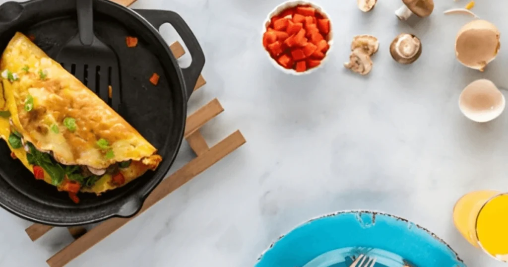 A skillet with a Greek omelette being cooked, surrounded by fresh ingredients like tomatoes, green onions, mushrooms, and feta cheese on a white countertop.