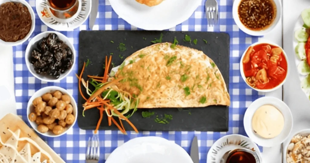 A full Mediterranean-style brunch spread with a Greek omelette as the centerpiece, surrounded by olives, bread, dips, and fresh vegetables on a blue-checkered tablecloth.